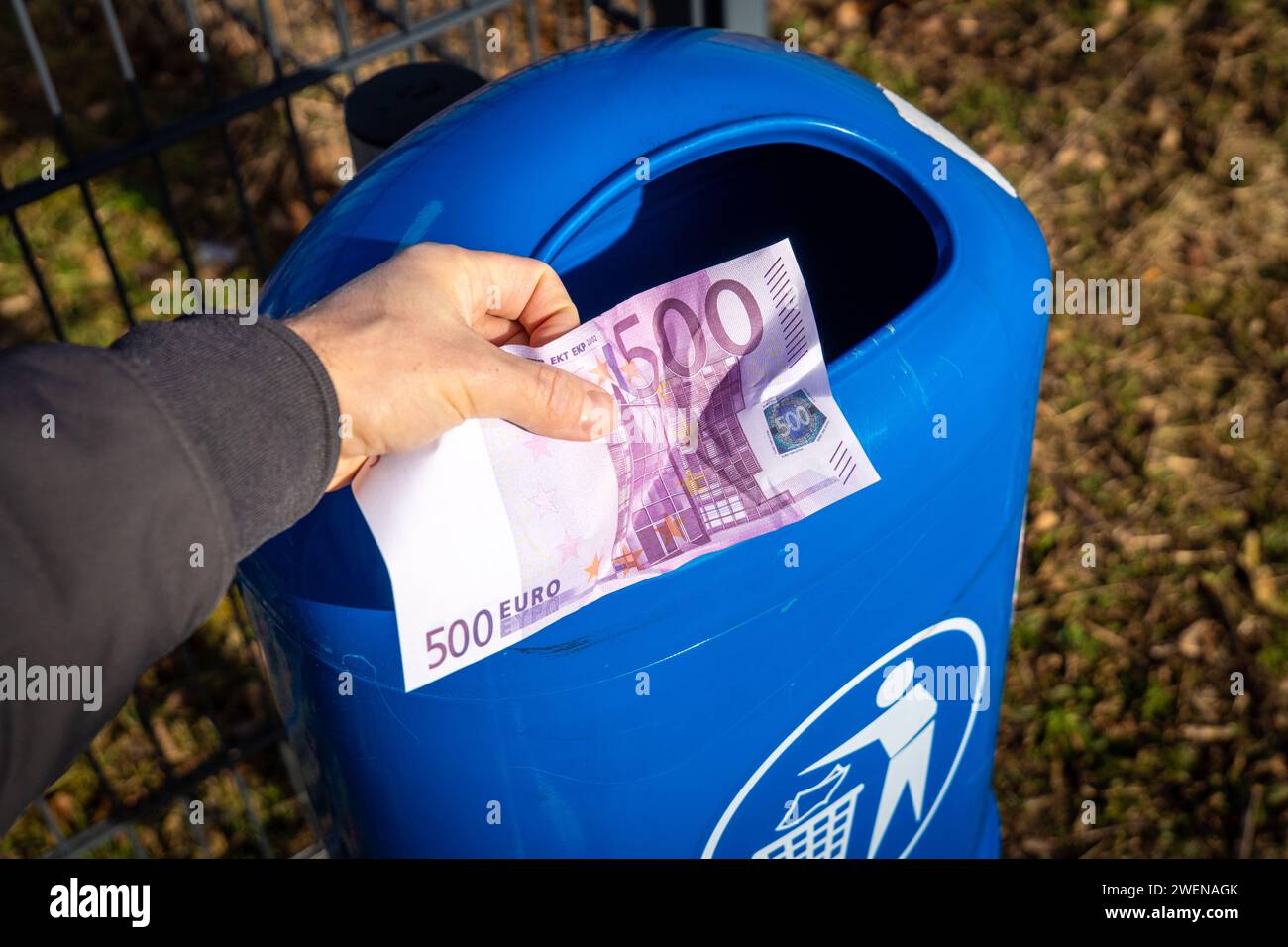 Langweid, Bavaria, Germany - January 26, 2024: Man throws euro cash banknotes into a blue trash can. Symbolic image of loss of value of money, recession and inflation *** Mann wirft Euro Bargeld Geldscheine in einen blauen Mülleimer. Symbolbild Wertverlust von Geld, Rezession und Inflation Stock Photo