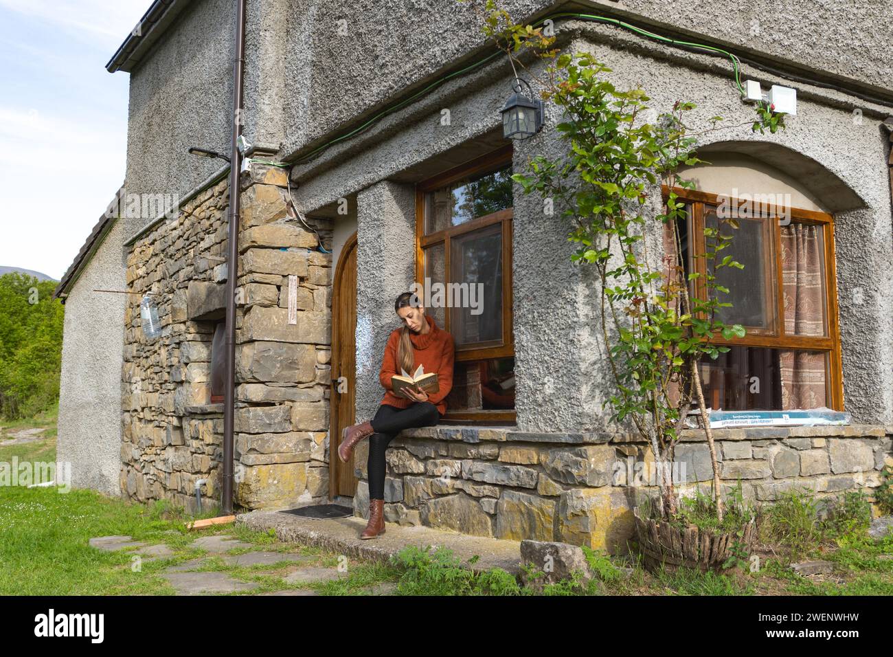Woman with crossed legs reads at the entrance of a country house, concentrated, turns the page Stock Photo
