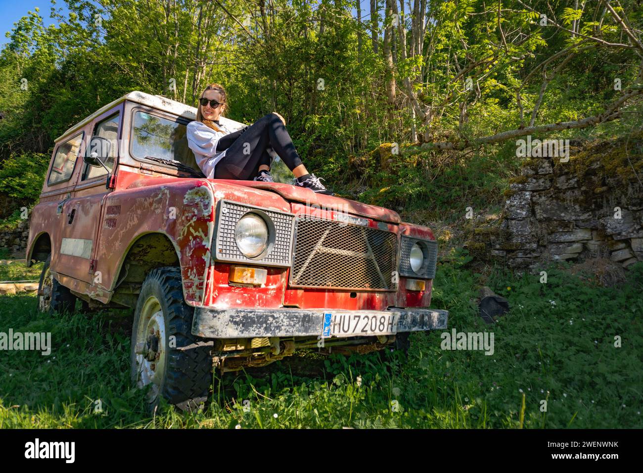 Woman sitting on the hood of a vintage red off-road parked in woods, sunbathing with sunglasses while smiling Stock Photo