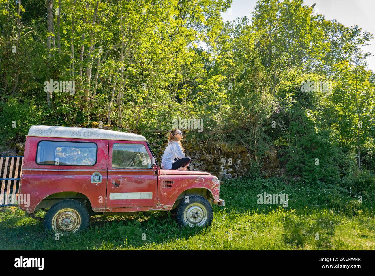 Woman sunbathing on the hood of a vintage red off-road in the forest, seen from the side while looking at the sky Stock Photo