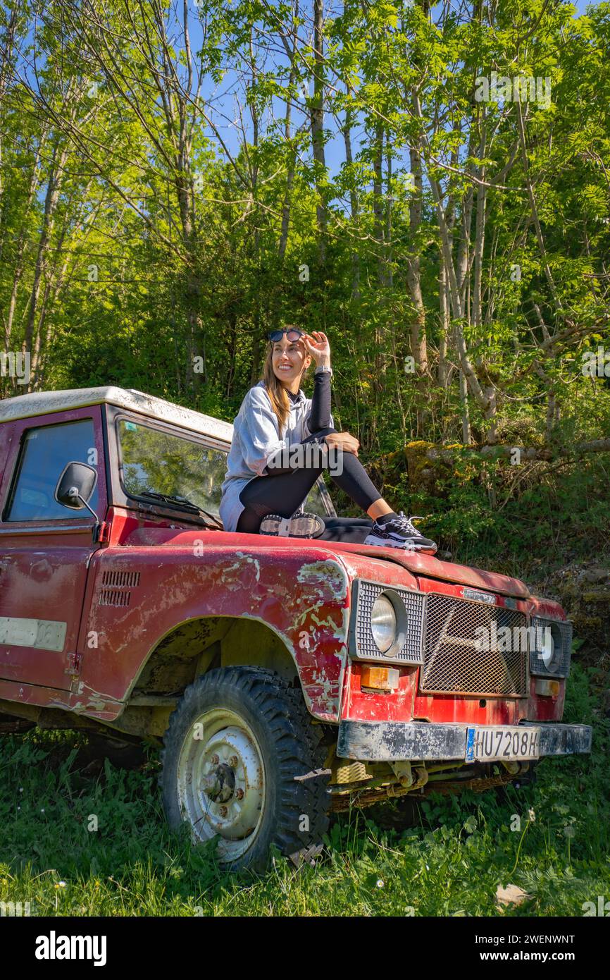 Woman sitting on the hood of a vintage red off-road parked in woods, takes off her glasses while smiling, vertical shot Stock Photo