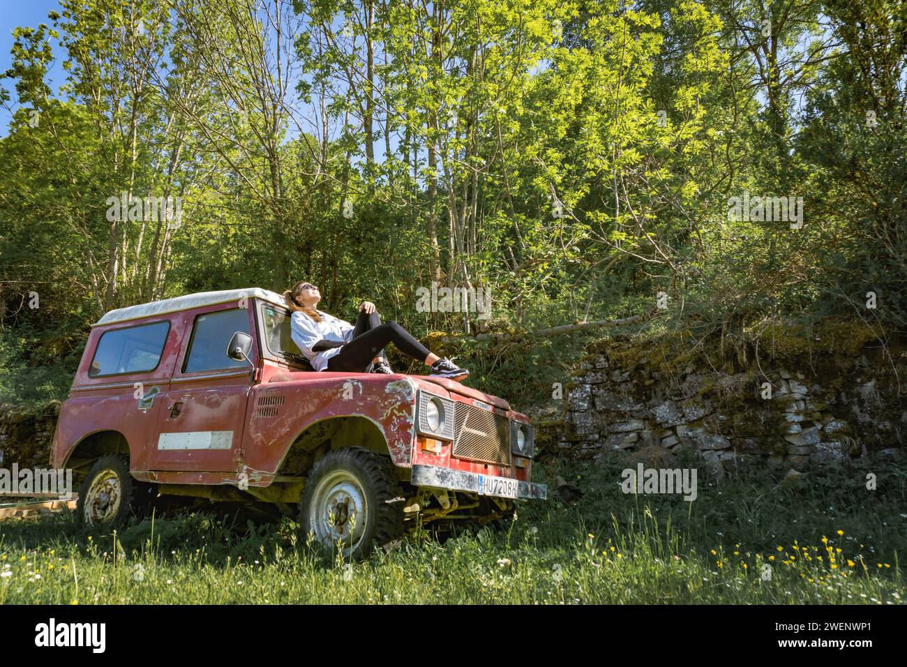 Woman sunbathing on the hood of a vintage red off-road vehicle in the forest, side view, relaxed Stock Photo