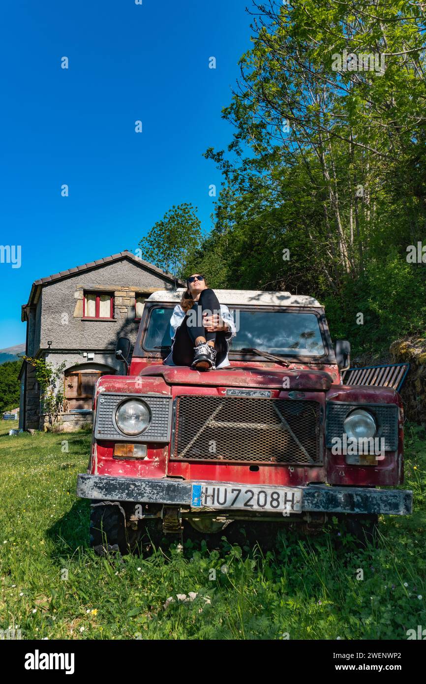 Woman sunbathing on the hood of a vintage red off-road vehicle in the forest, front view, vertical shot Stock Photo