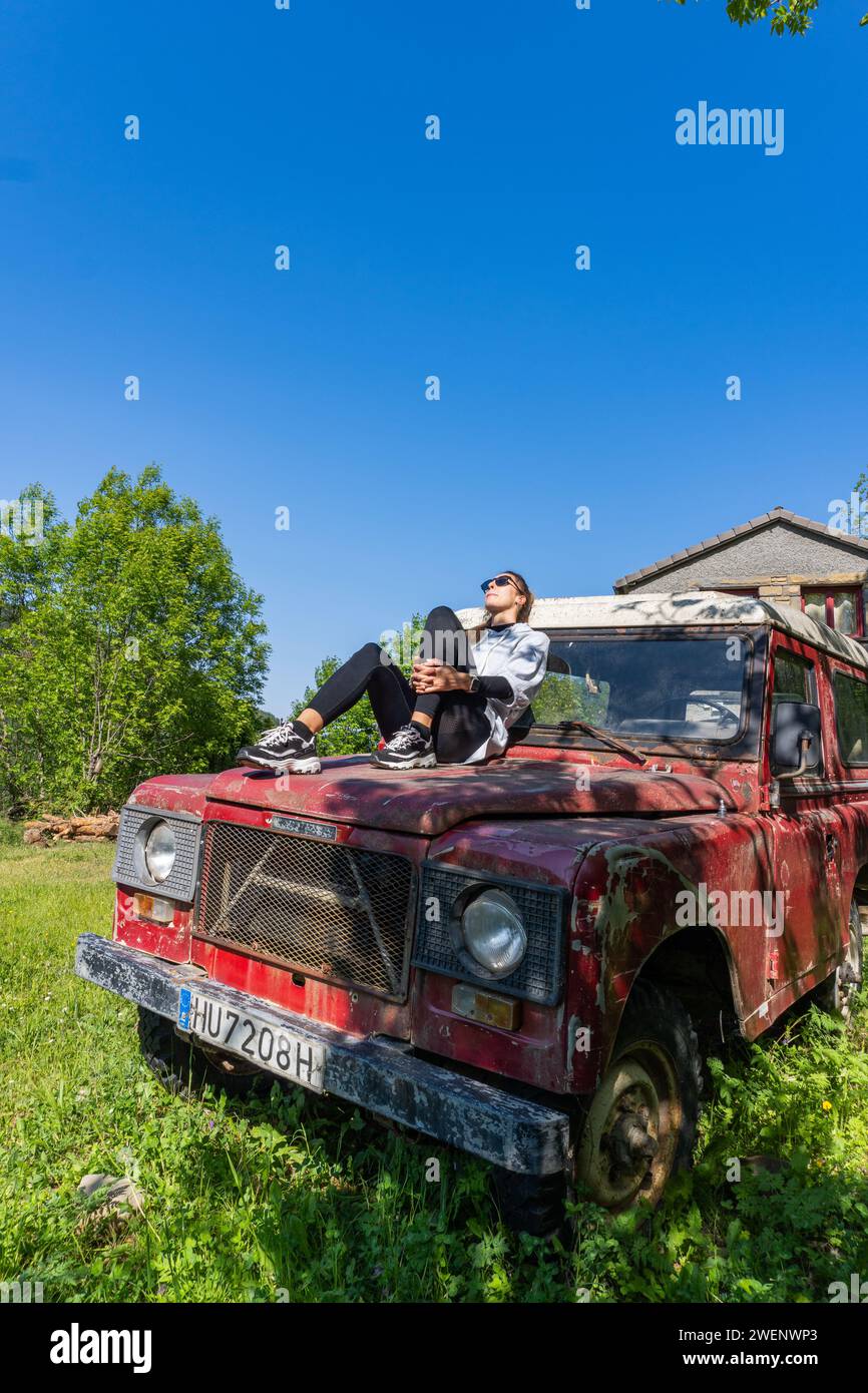 Woman sunbathing on the hood of a vintage red off-road vehicle in the forest, side view, vertical shot Stock Photo