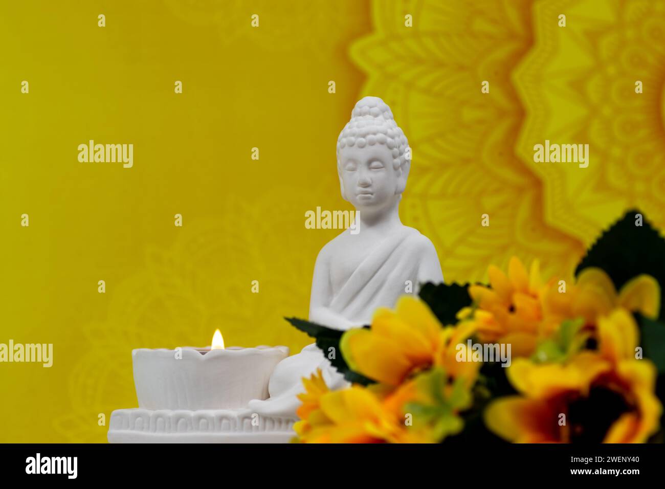 Close up of white marble figurine of Siddhartha Gautama, known as Buddha, with candle burning and decorative flowers, colorful thematic background Stock Photo