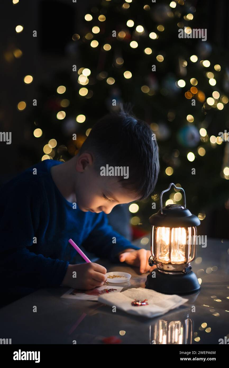 Little boy making handmade decoration for Christmas tree at night. Christmas tree with lights on background. Winter holidays concept Stock Photo