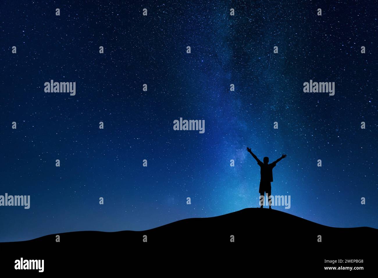 Beautiful night landscape of the milky way in the cloudy sky and silhouette of a young guy with his hands up. Victory and success concept. Stock Photo