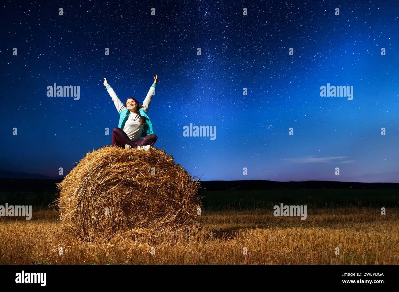 Young happy girl with raised arms sits on a round haystack in a field against the background of the starry sky. Night landscape of the starry sky with Stock Photo