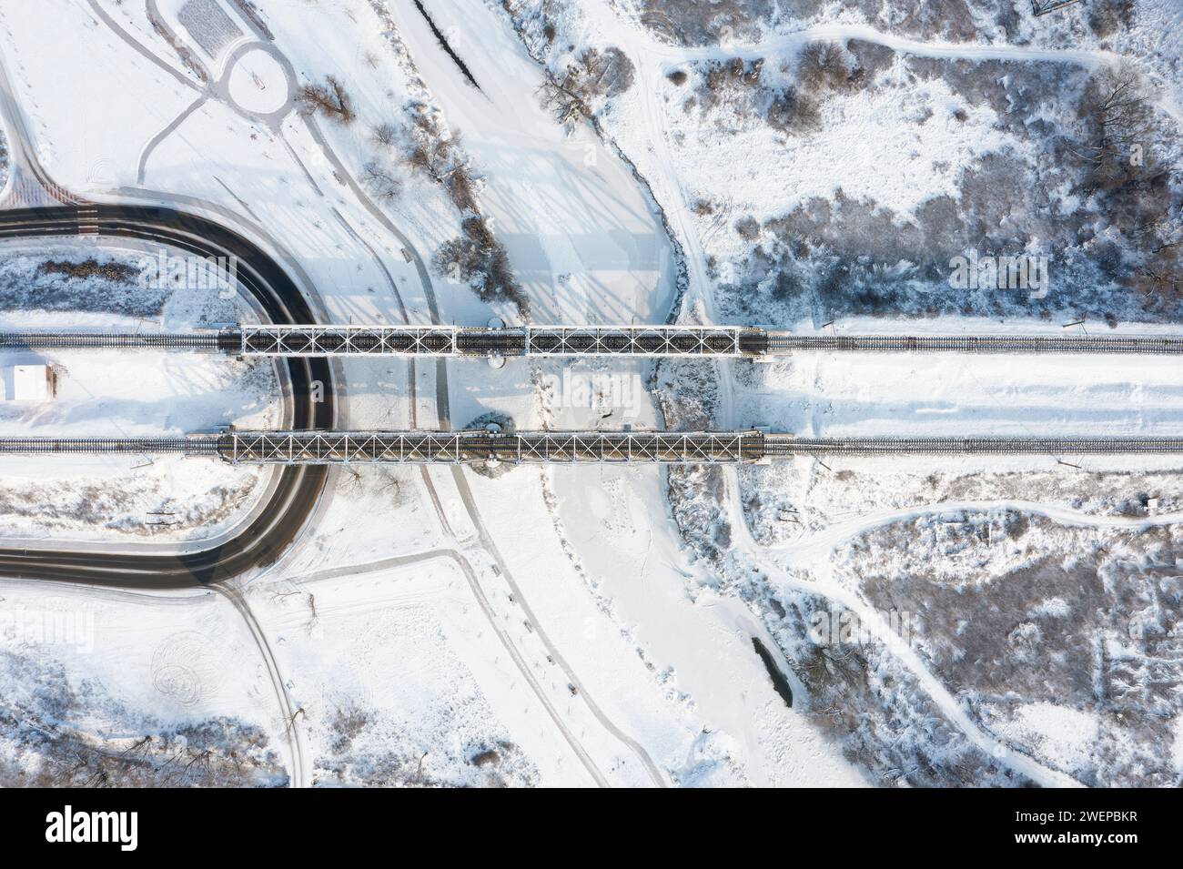 Top aerial view of the railway bridge over the frozen river covered with snow on a winter sunny day. Stock Photo