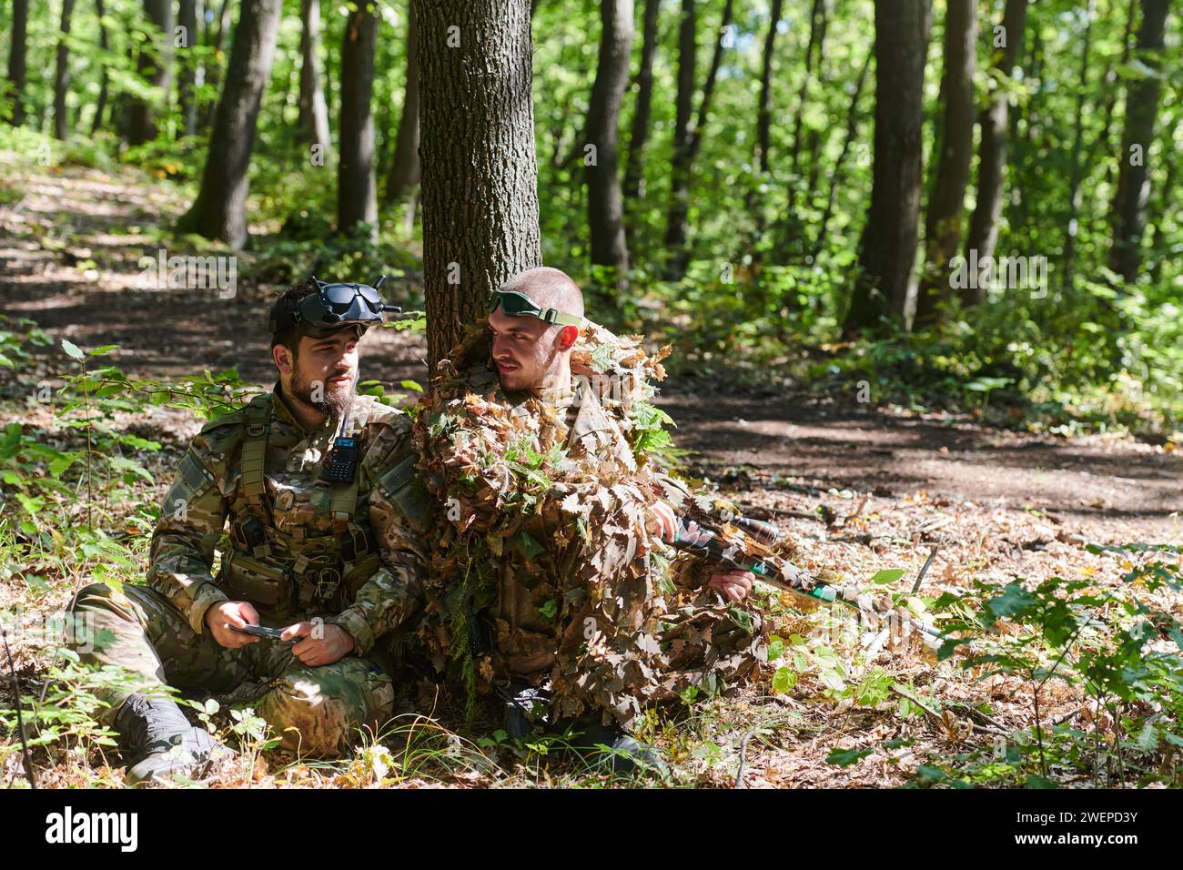 A skilled sniper and a soldier operating a drone with VR goggles strategize and observe the military action while concealed in the forest Stock Photo