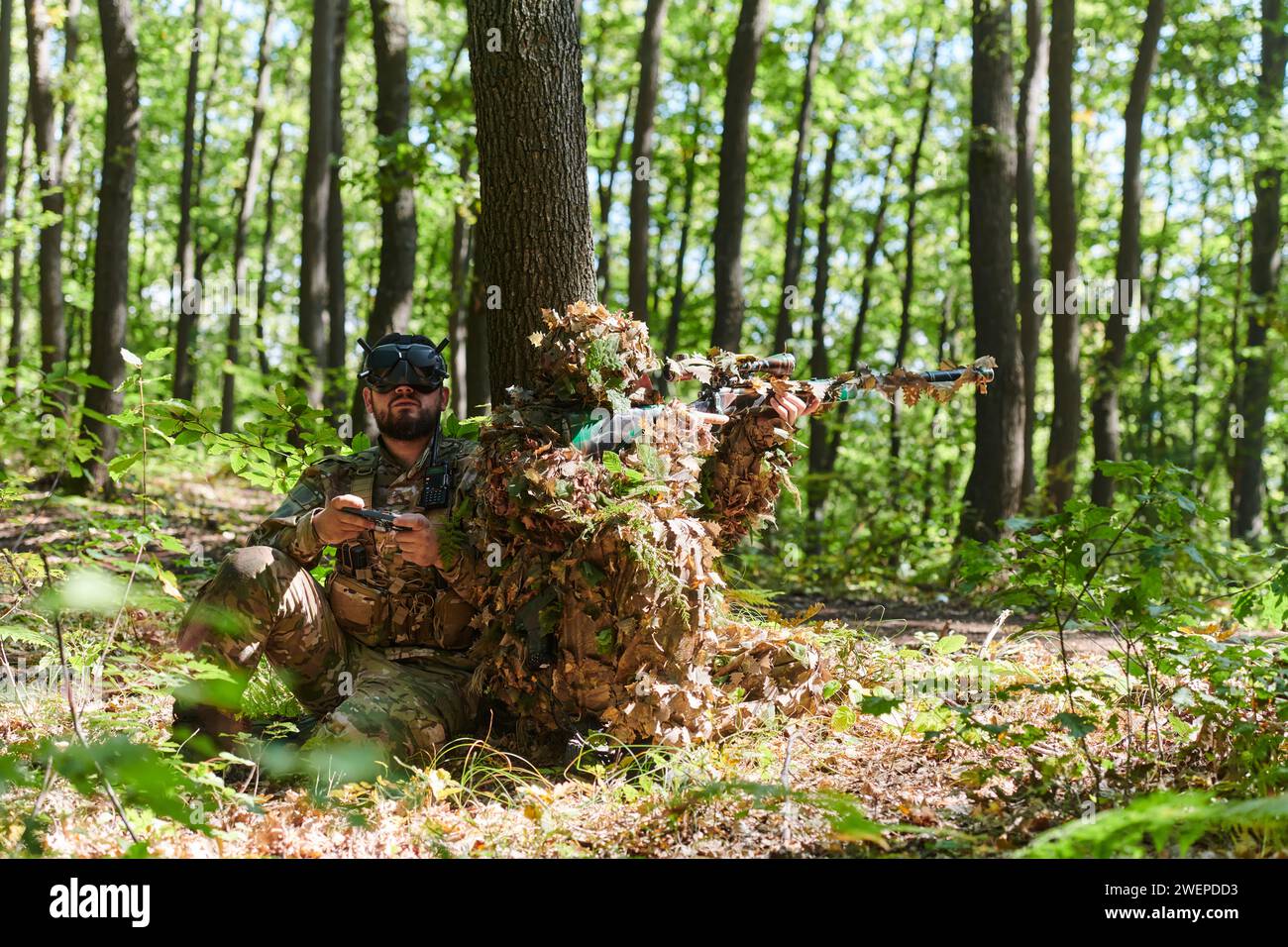 A skilled sniper and a soldier operating a drone with VR goggles strategize and observe the military action while concealed in the forest Stock Photo