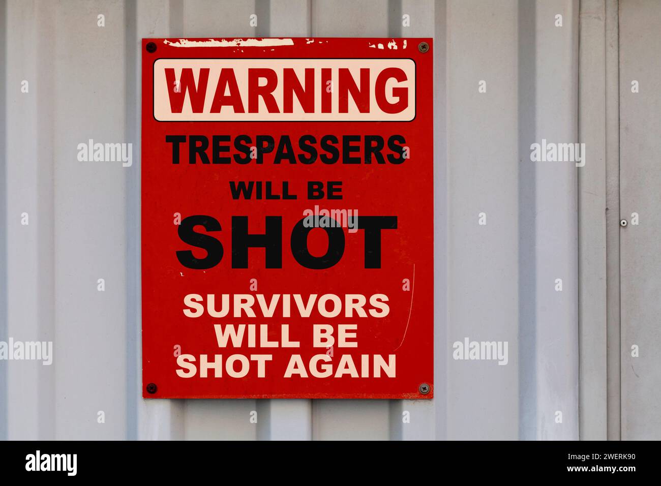 Warning sign drawn on a dark red placard with written in 'Warning - Trespassers will be shot, survivors will be shot again'. Stock Photo