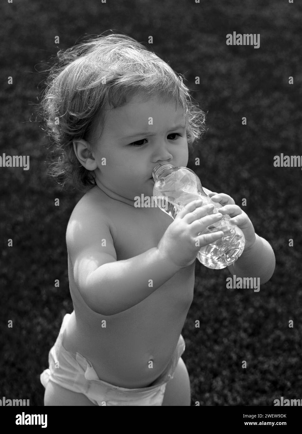 Baby drinking water. Close-up of little blonde child drinking fresh and pure water from bottle with a blurred green grass background. Stock Photo