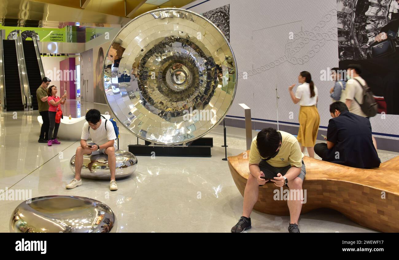 People sit near a silver coloured loudspeaker shaped display in Siam Mall or Siam Paragon shopping centre, a large mall in central Bangkok, Thailand, Southeast Asia Stock Photo