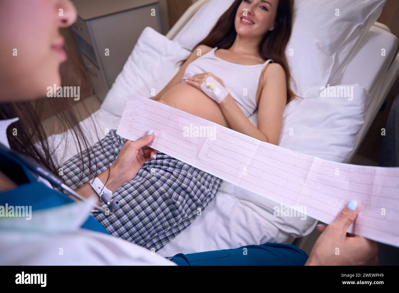 Obscure face of female nurse checking electrocardiogram of young caucasian pregnant woman on medical bed in hospital. Concept of pregnancy and materni Stock Photo