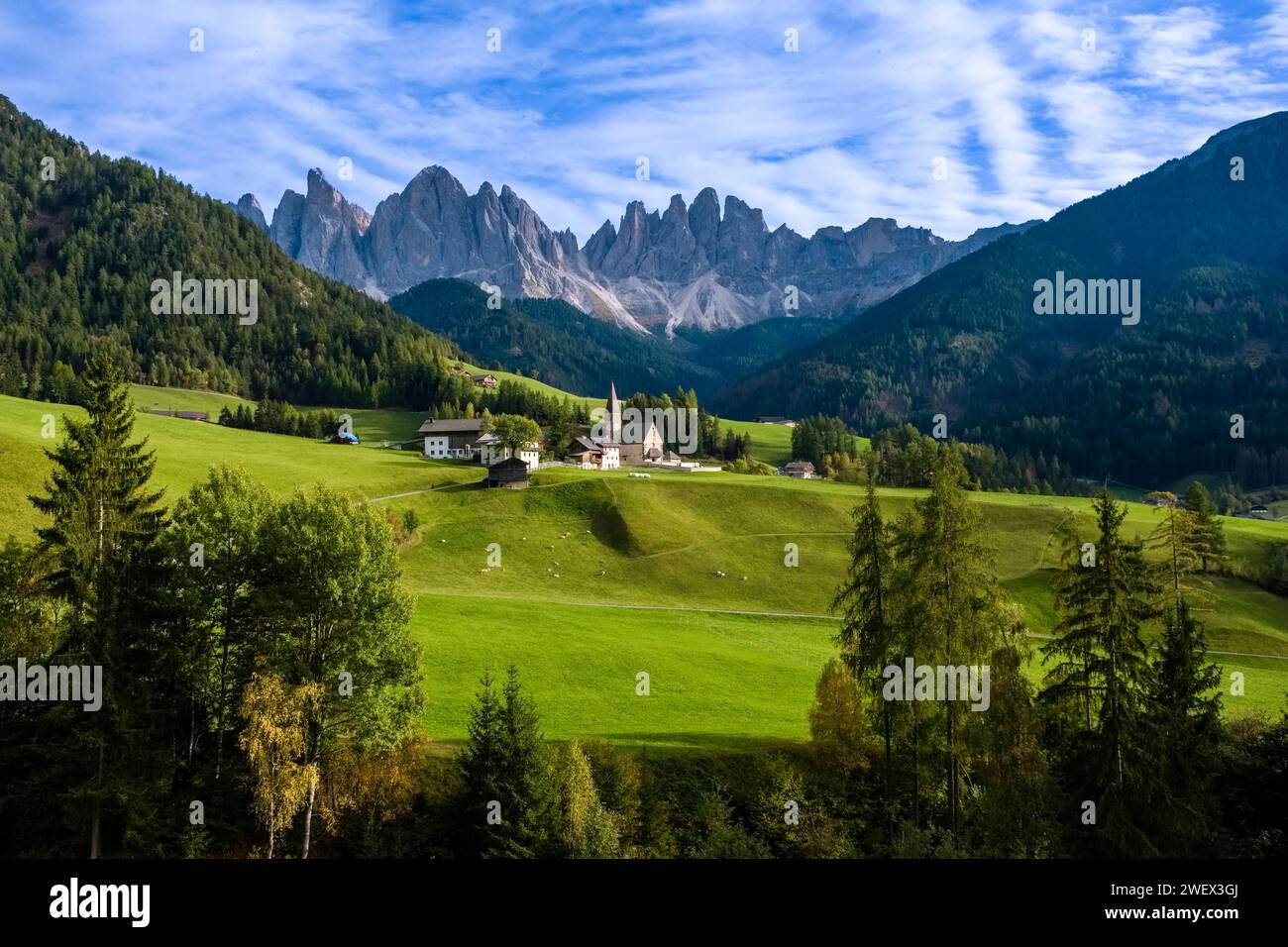 View of the Church of St. Magdalena in Villnöss valley, north faces and summits of Odle group in the distance, in autumn. Bolzano Trentino-Alto Adige Stock Photo