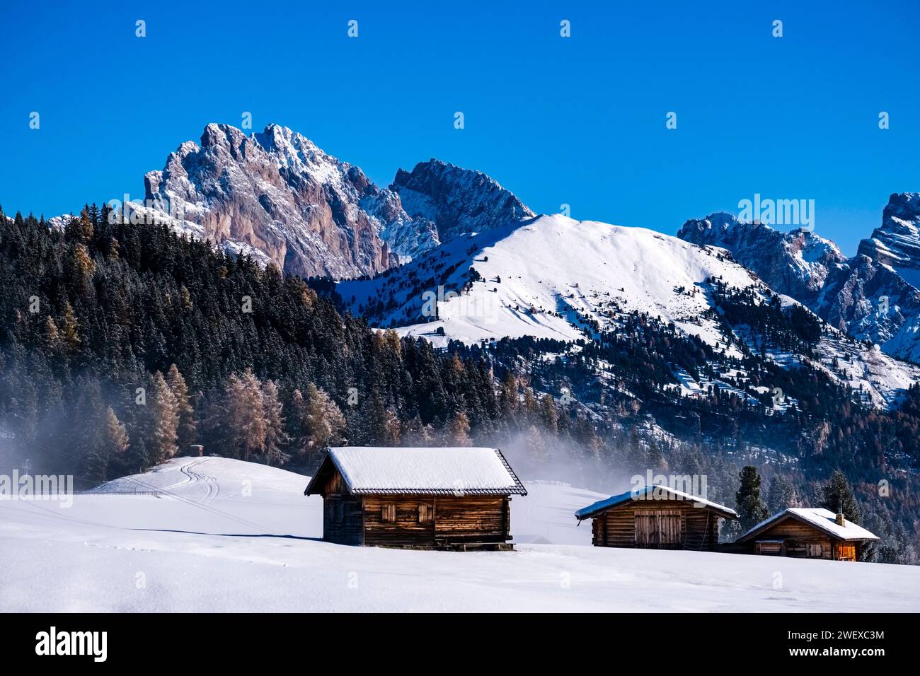 Hilly agricultural countryside with wooden huts and snow-covered pastures at Seiser Alm, in winter, summits of Odle group in the distance. Kastelruth Stock Photo