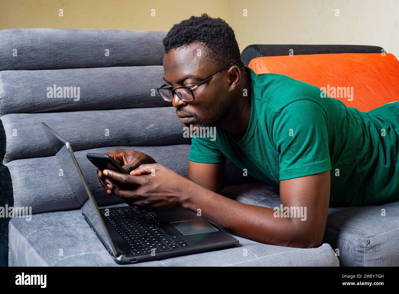 young man wearing optical glasses lying on sofa at home uses mobile phone and laptop. Stock Photo