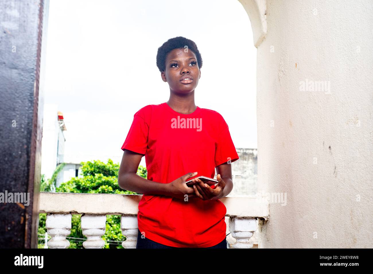 beautiful young girl wearing red t-shirt standing on the balcony of the house with a mobile phone in hands. Stock Photo