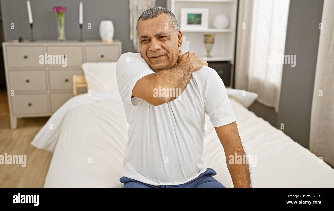 Mature hispanic man in bedroom setting displaying discomfort or soreness, holding his neck. Stock Photo
