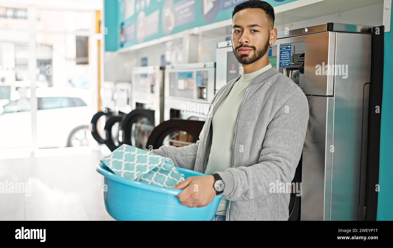 Young hispanic man holding basket with clothes at laundry facility Stock Photo
