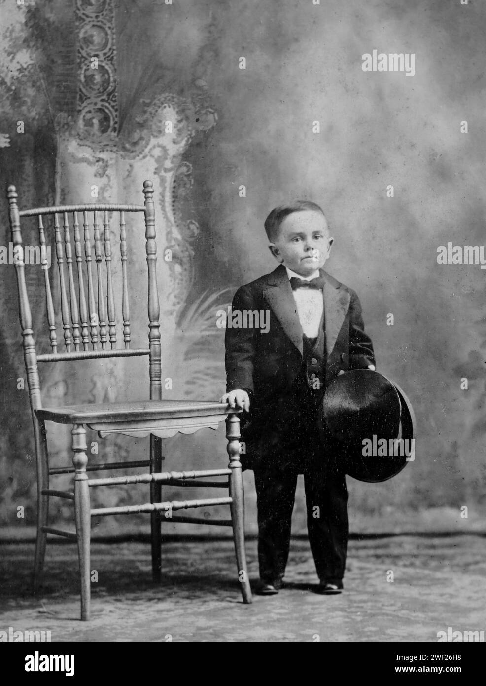 Nineteen year old little person man poses by a chair in a studio portrait, ca. 1895. Stock Photo