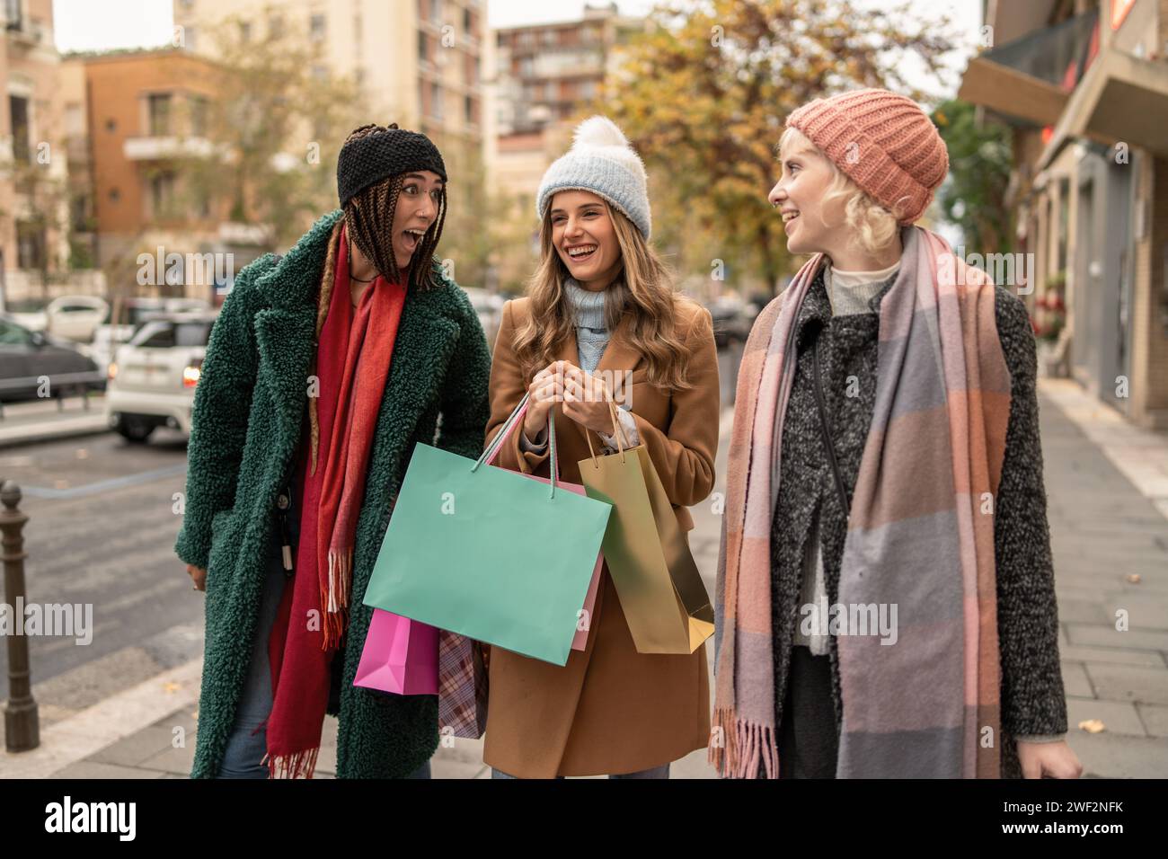 Three young women stroll through city streets, smiling and chatting happily with numerous colorful shopping bags in hand, immersed in the vibrant autu Stock Photo