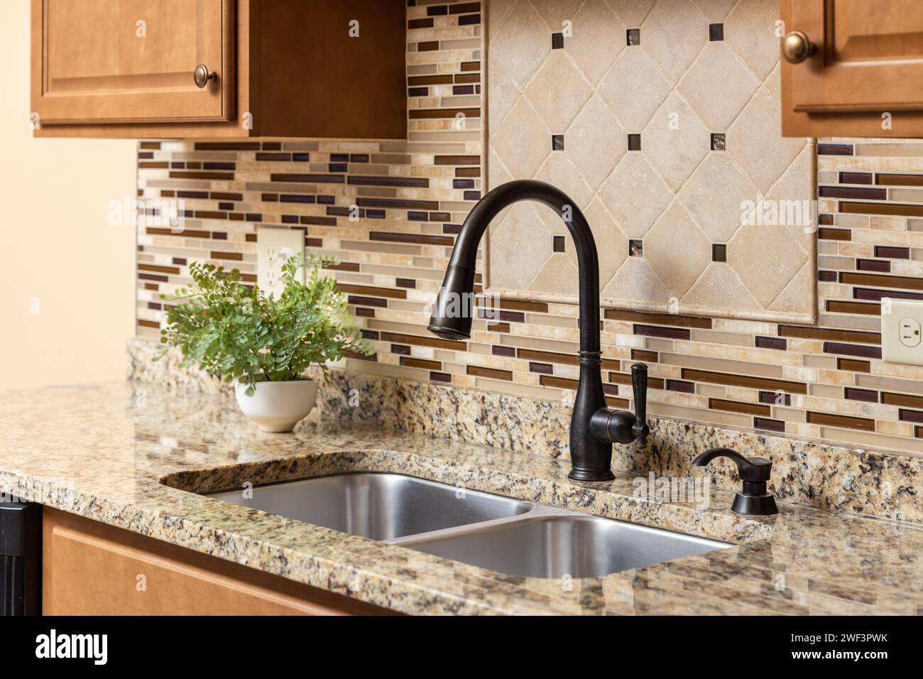 A kitchen faucet detail with wood cabinets, an oil rubbed bronze faucet, quartz countertop, and glass and stone tile backsplash. Stock Photo