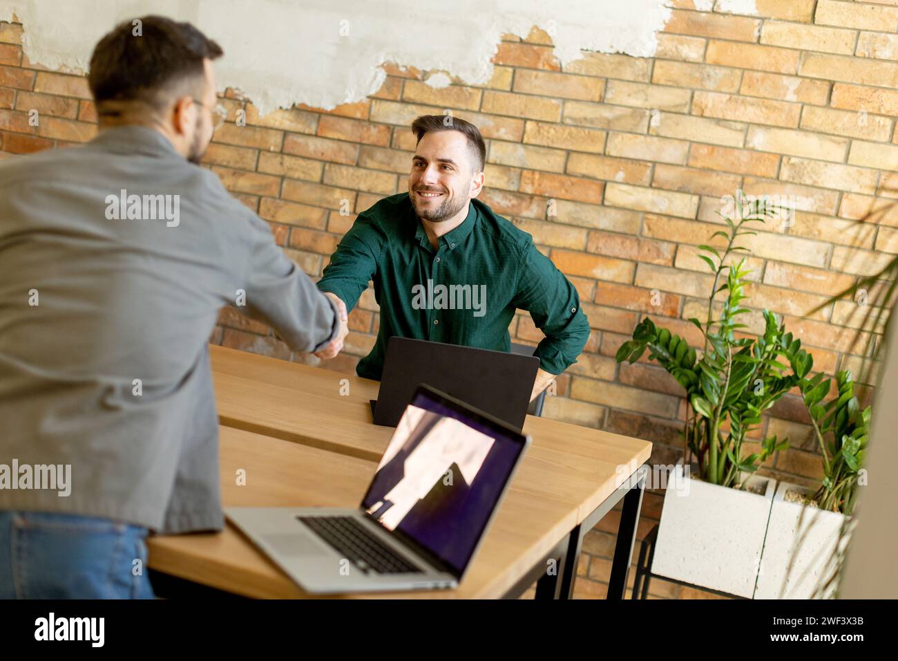 Two professionals engage in a welcoming handshake across a wooden table adorned with laptops, signaling a successful meeting or partnership in a conte Stock Photo