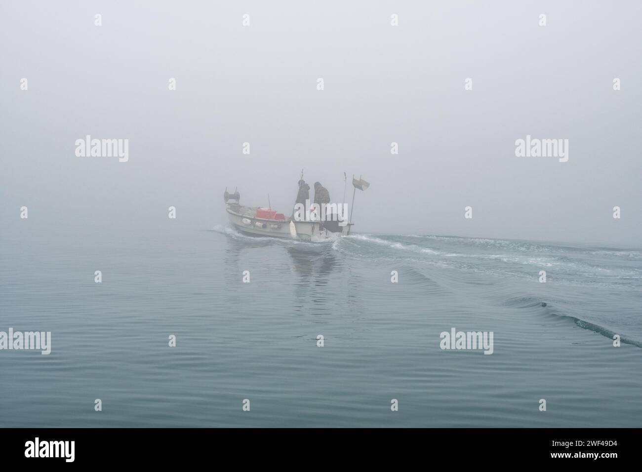 Grado, Italy - January 28th, 2024: a small fishing boat with two fishermen departs from the Mandracchio port for a fishing trip on a day of thick fog. Stock Photo