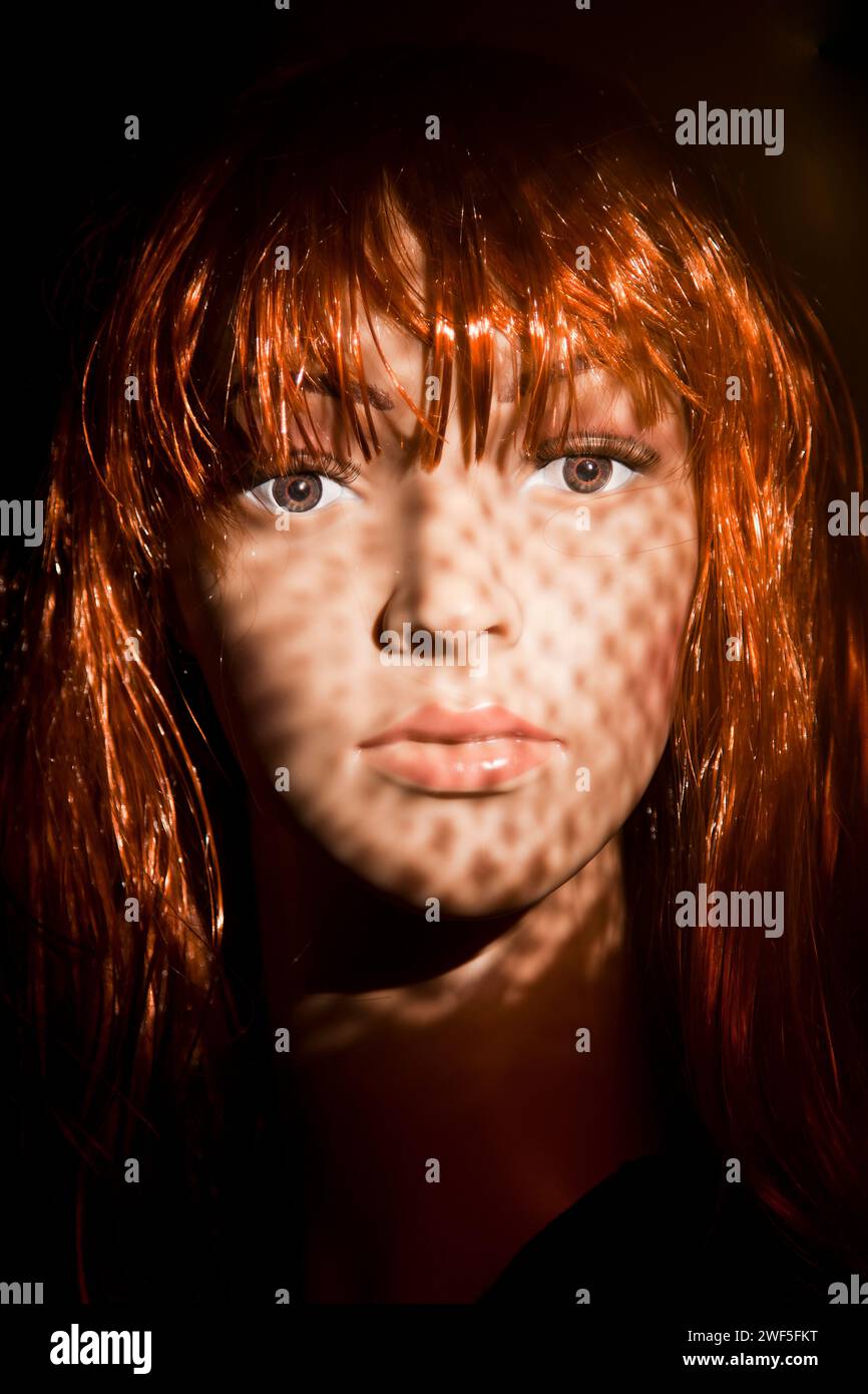 Red haired plastic woman mannequin with bright hair posing on a black background with shadow dots projected on her face as a special effect Stock Photo