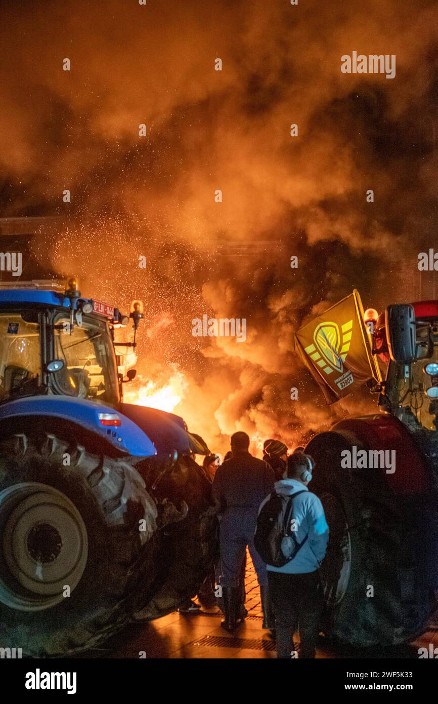 Turnhout, Antwerp, Belgium, 28th of January, 2024, The image captures an evocative scene from a farmers' protest against the Nitrogen Agreement in Turnhout, Belgium. Amidst the night, the fervent glow of a bonfire sets the stage, with thick smoke billowing into the dark sky. The tractor, a symbol of the farming profession, is positioned prominently, as a group of protestors gathers, some holding flags representative of their cause. The intense and spirited atmosphere is palpable, with the dramatic illumination reflecting the critical nature of the protest. Nighttime Vigil of Farmers' Protest i Stock Photo