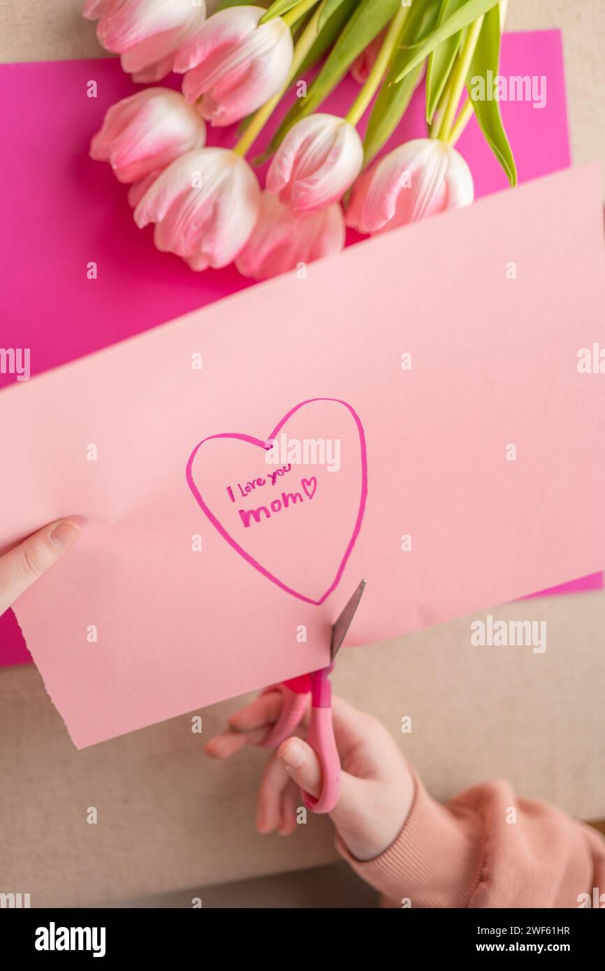 Mothers Day. DIY mom card.Child cuts a heart out of paper at the table. child makes a card for his mother.Flowers and cards for mom. Stock Photo