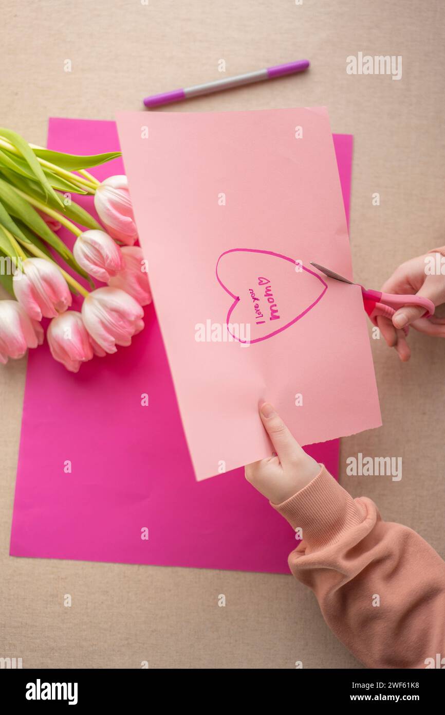 Mothers Day. Child cuts a heart out of paper at the table.child makes a card for his mother.Flowers and cards for mom.DIY mom card. Stock Photo