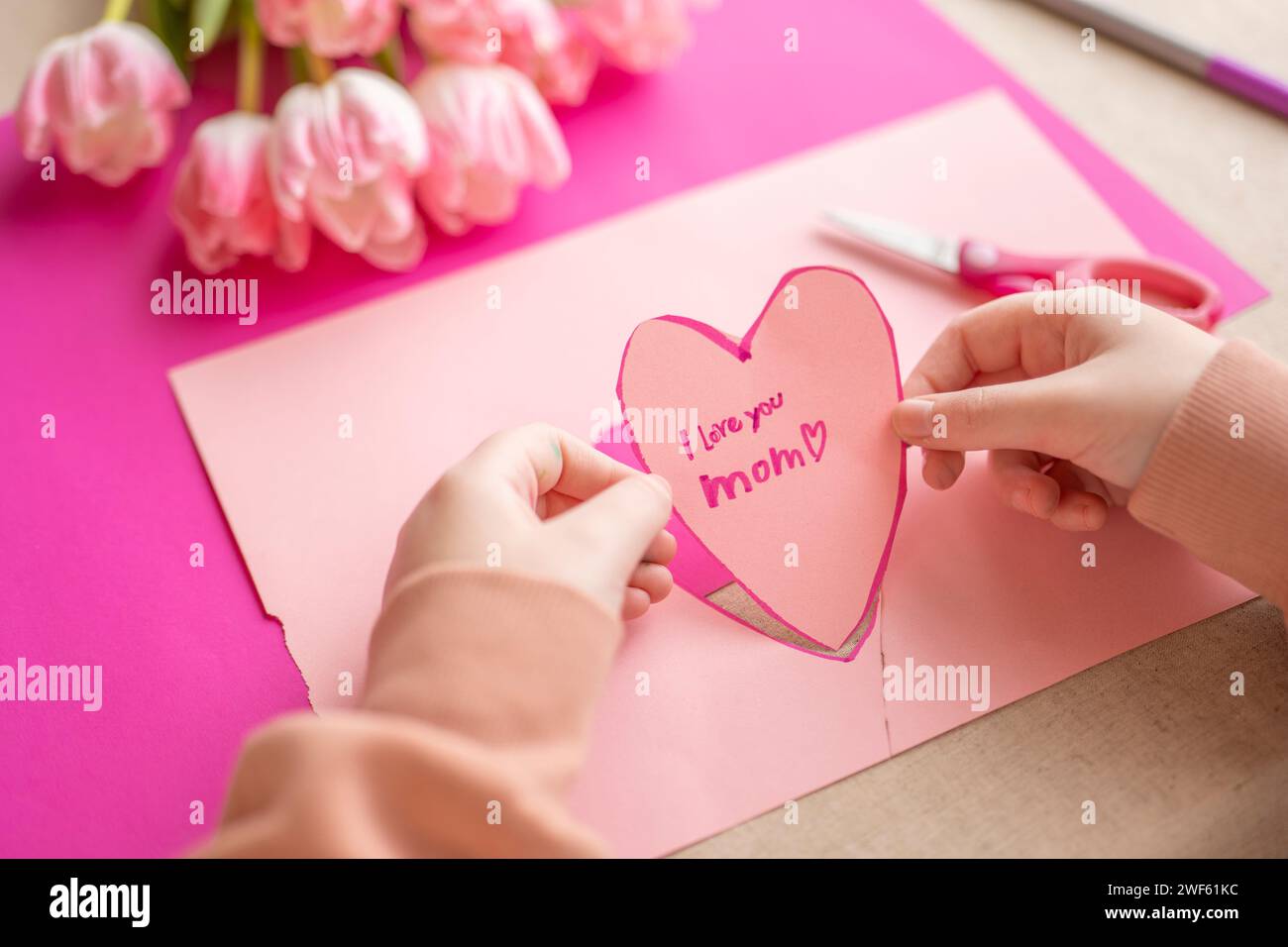 Mothers Day.I love you, Mom.Flowers and cards for mom. Childrens hands holding a heart card.Pink heart card and pink tulips.child makes a card for his Stock Photo