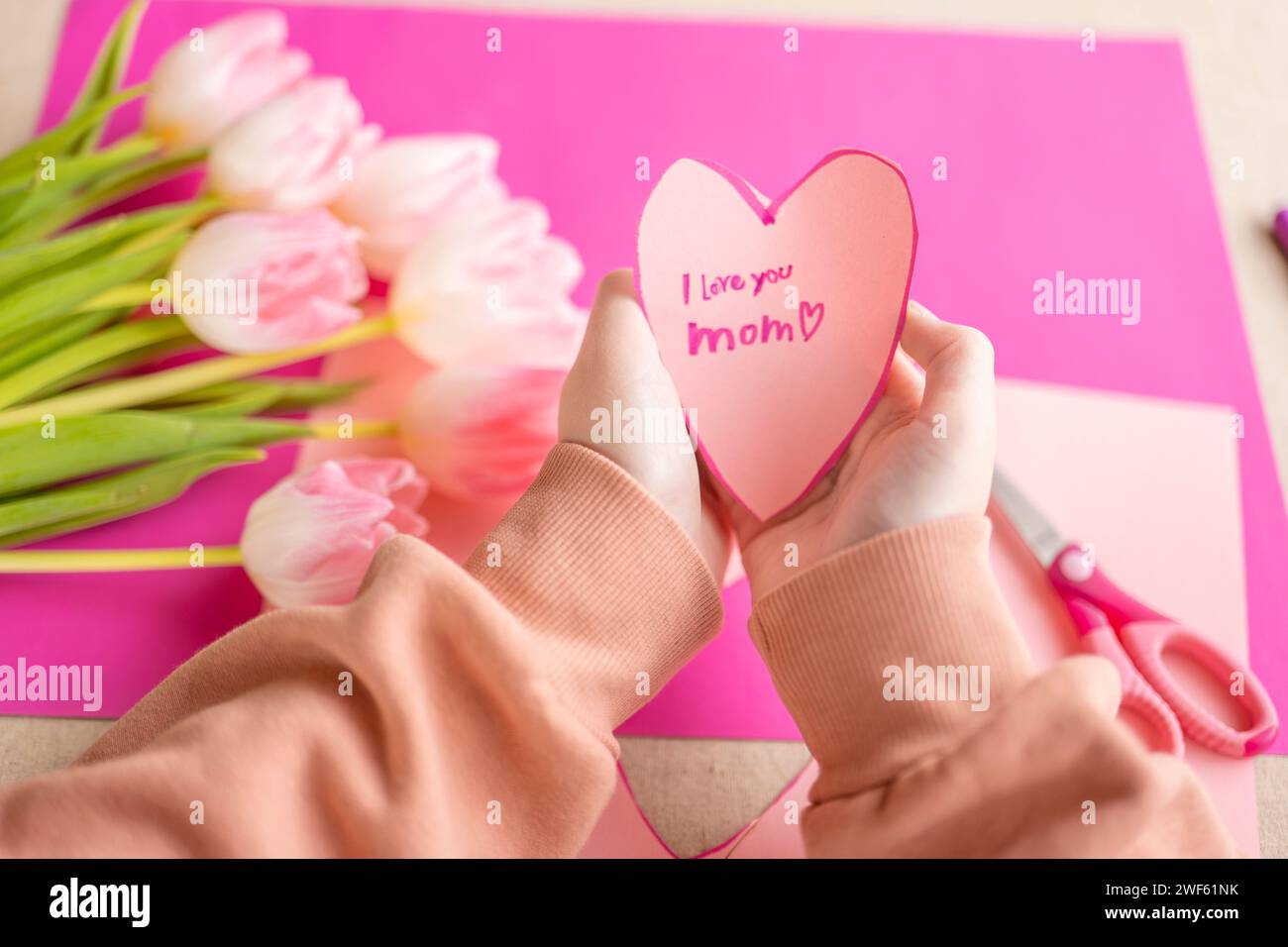 Mothers Day.I love you, Mom. Childrens hands holding a heart card.Pink heart card and pink tulips.child makes a card for his mother.Flowers and cards Stock Photo