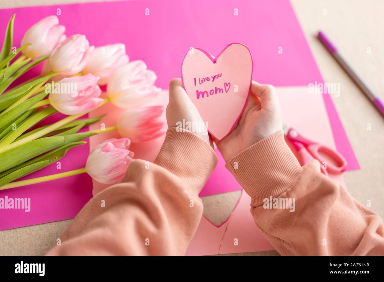 I love you, Mom. Childrens hands holding a heart card.Pink heart card and pink tulips.child makes a card for his mother.Flowers and cards for mom.moms Stock Photo