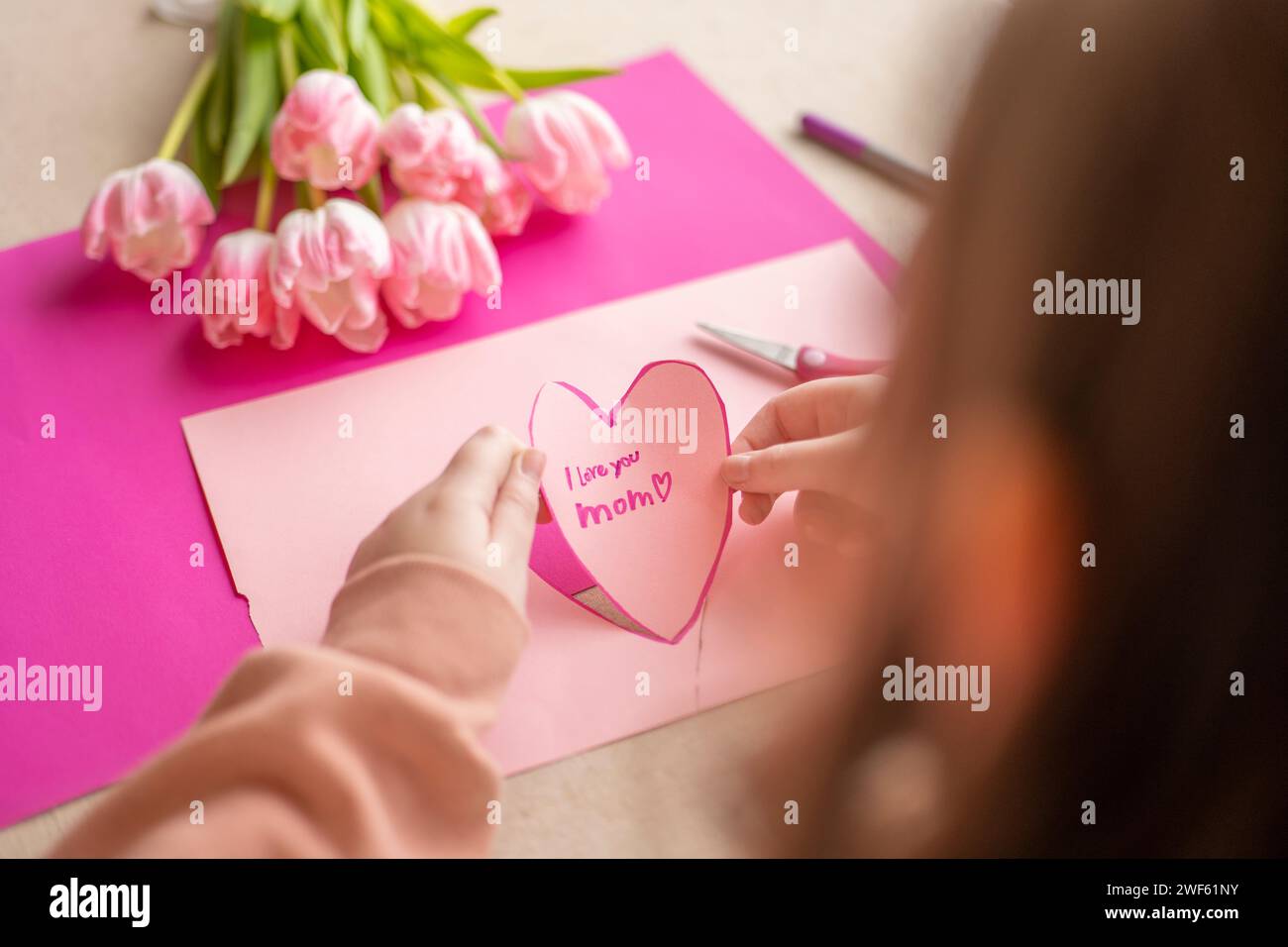 moms day. I love you, Mom. Childrens hands holding a heart card.Pink heart card and pink tulips.child makes a card for his mother. Stock Photo