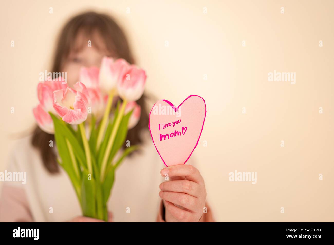 Mothers Day. daughter with flowers and card on a beige background.Laughing girl with tulips and heart card.Pink heart card and pink tulips close-up in Stock Photo