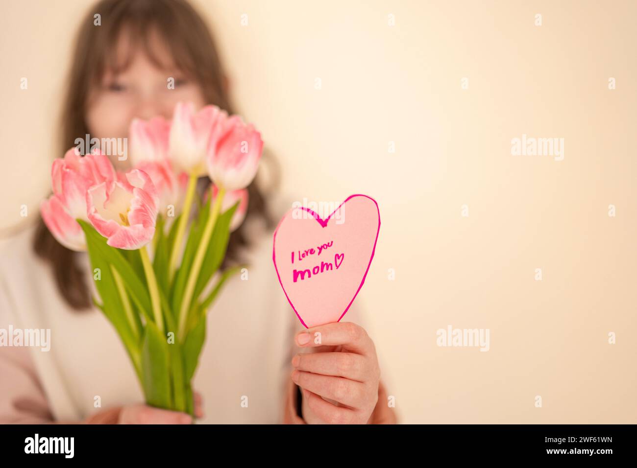 Mothers Day.Smiling daughter with flowers and card on a beige background.Laughing girl with tulips and heart card.Pink heart card and pink tulips Stock Photo