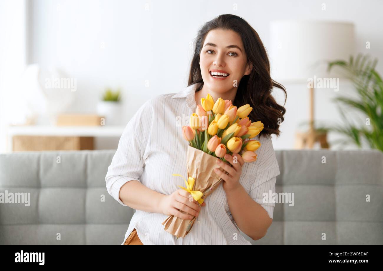 Beautiful young woman with flowers tulips in hands sitting on sofa at home. Stock Photo