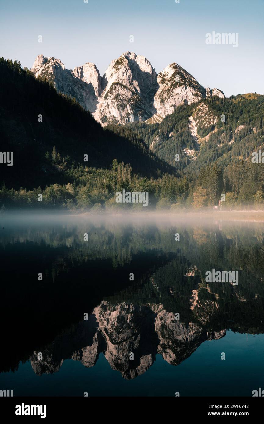 A scenic misty mountain range with trees reflecting on calm waters. Stock Photo