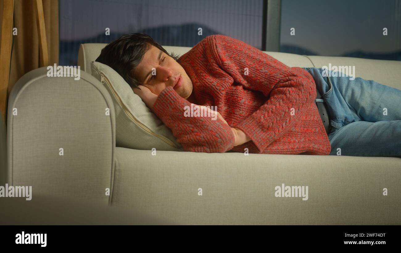 man lays down himself on pillow on the sofa in living room. Male trying to relax after tiring work day at home Stock Photo