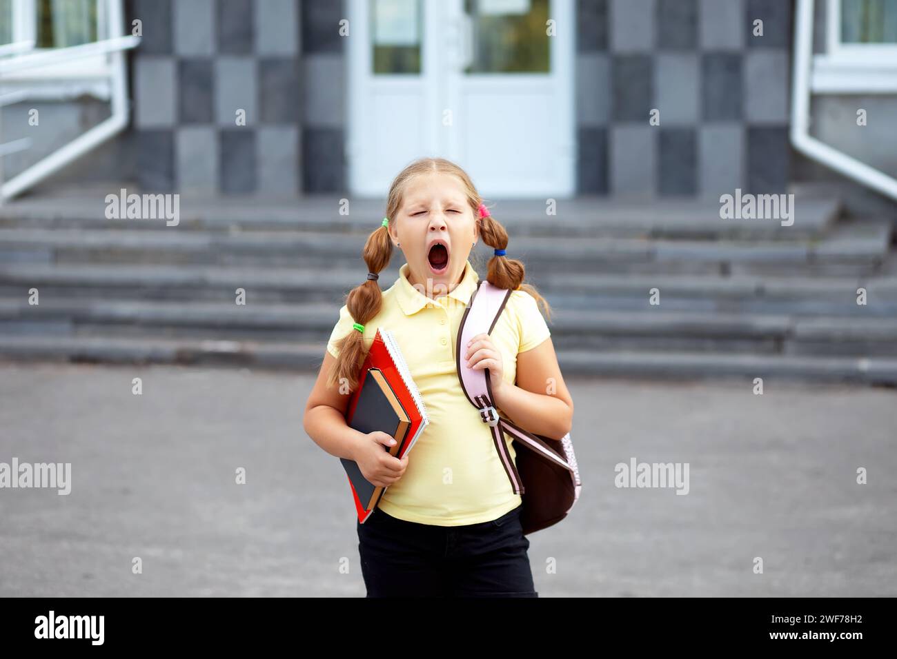 Pretty schoolgirl yawning after lessons on the doorstep of the school. Tired student - hard school day Stock Photo