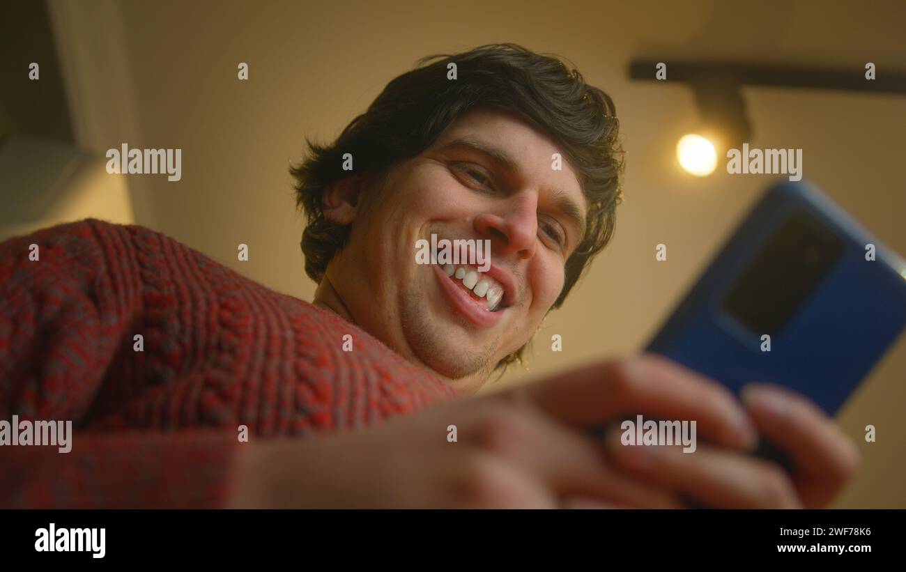 Low angle shot of smiling young man standing in living room typing text message with his smartphone. Male rejoices at the message on his smartphone fe Stock Photo