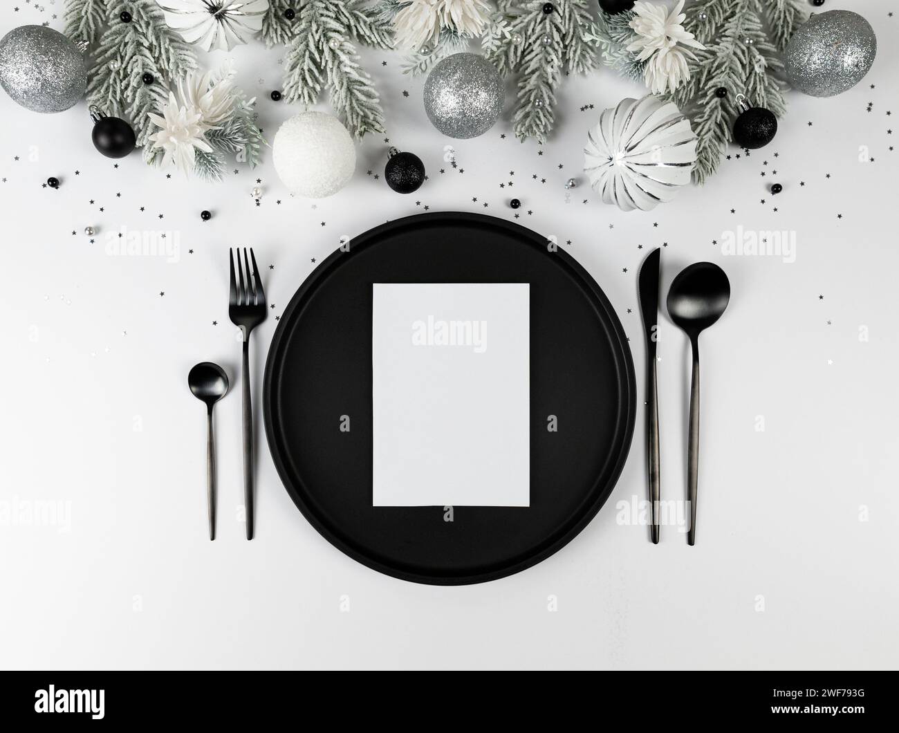 Christmas composition, white and silver decorations, black plate and cutlery, fir tree branches, white card on white background. Stock Photo