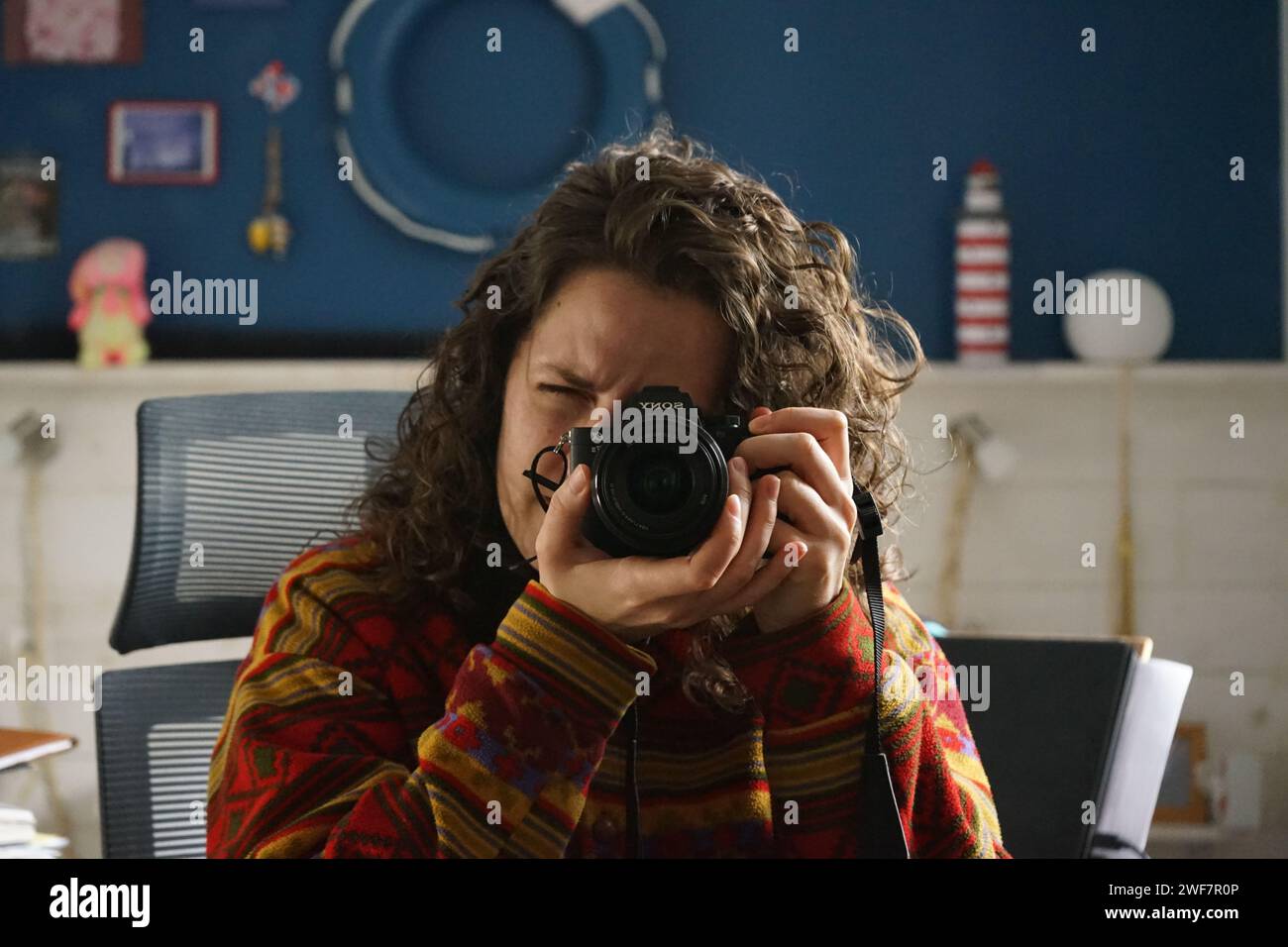Young white woman with curly hair taking a picture of herself from a sony alpha 7 ii mirrorless camera in a home office with blue and white wall Stock Photo