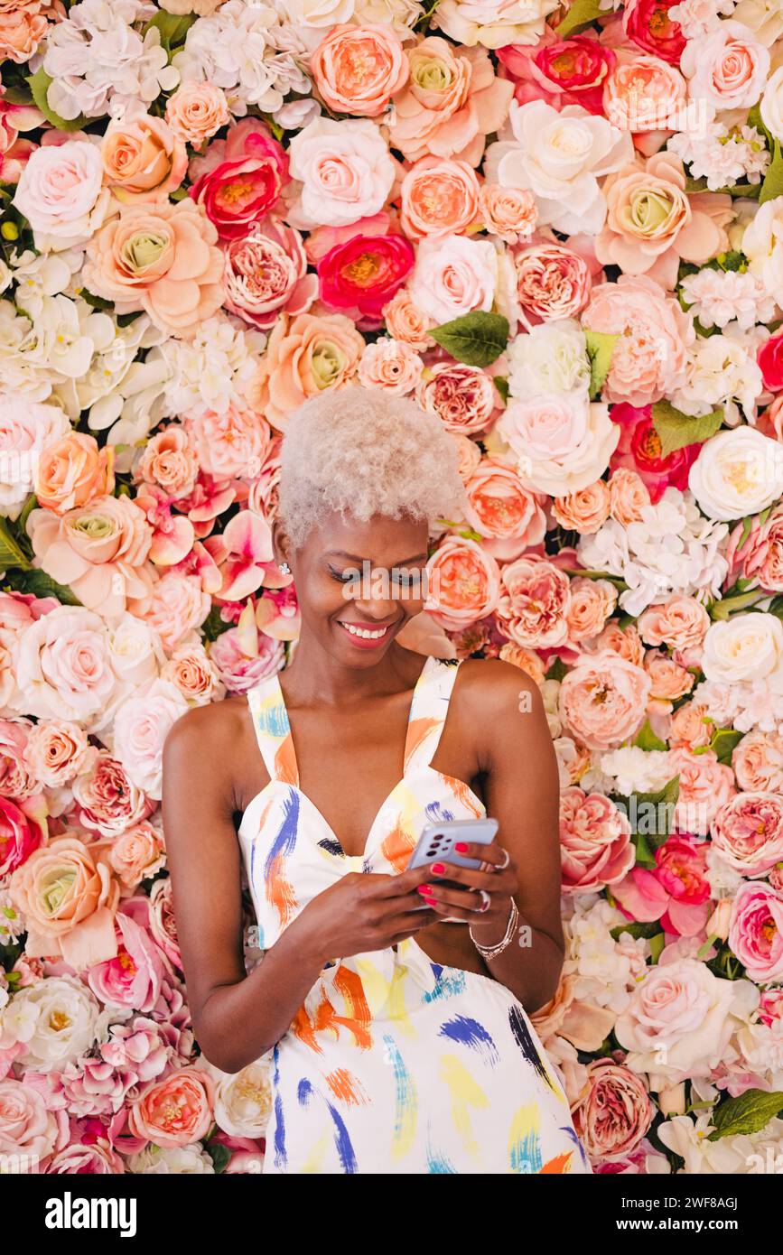 Portrait of positive young panamanian woman with short blond hair in stylish dress. She is smiling and besides a blooming flowers wall with pink tones Stock Photo