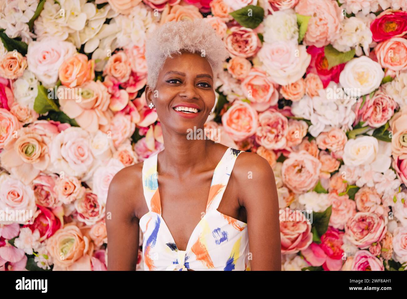 Positive panamanian woman with dyed hair in stylish outfit smiling and looking at camera while standing near blooming flowers Stock Photo