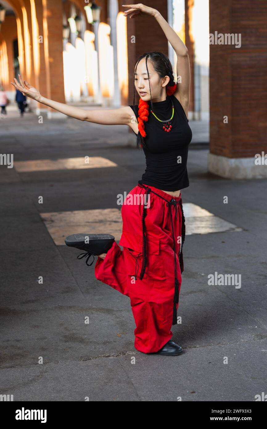 An urban dancer in striking red trousers performs an elegant pose inspired by oriental dance on a city walkway. Stock Photo